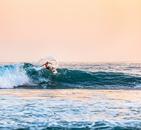 Male surfer riding a turquoise wave under a hazy peach sunset.