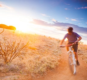 Man riding a bike on a desert trail under an intense, orange sunset.
