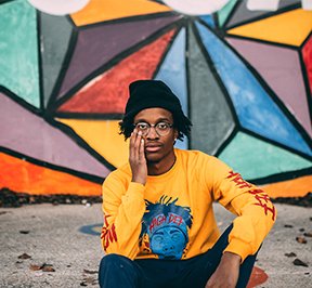 Young, black man sitting in front of a colorful mural. 